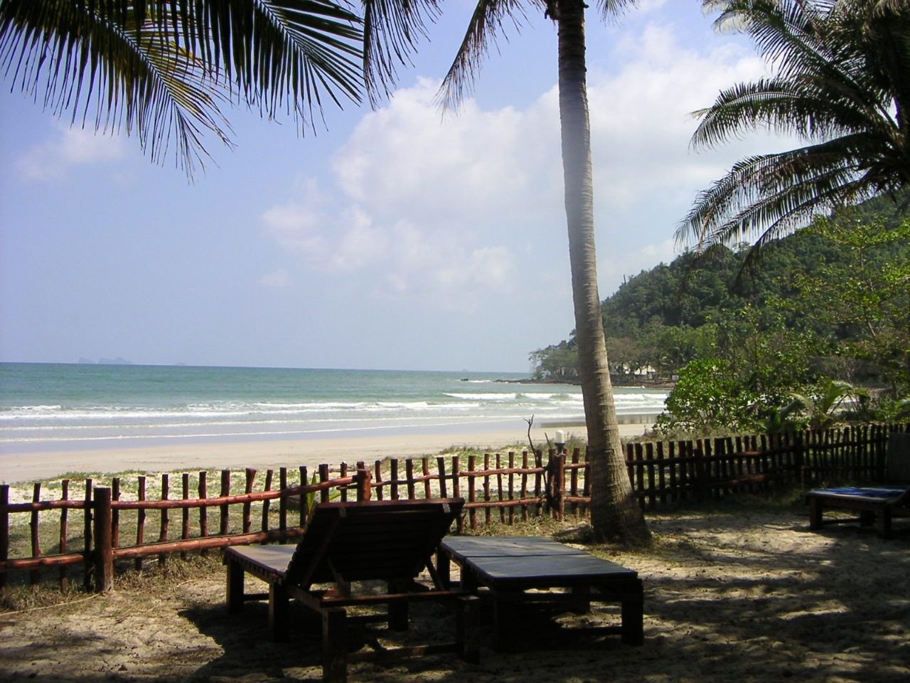 Une plage tropicale. On y voit du sable, l'océan avec des vagues, des palmiers, une clôture en bois et des chaises longues, suggérant un cadre de détente ou de vacances, photo gratuite