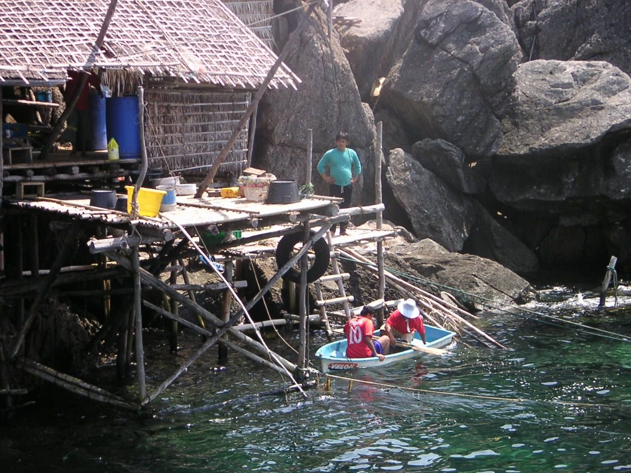 A wooden structure with a thatched roof built over the water, and people, fishermen, in a small boat below, with large rock formations in the background, free photo