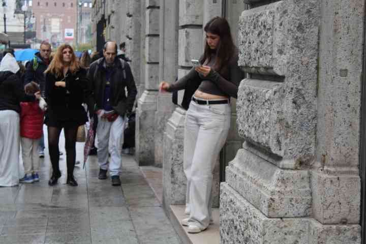 Une scène de rue urbaine où l'on peut observer plusieurs personnes et des éléments architecturaux, une femme est adossée à un mur en pierre, regardant son téléphone portable, d'autres personnes marchent sur le trottoir, photo gratuite