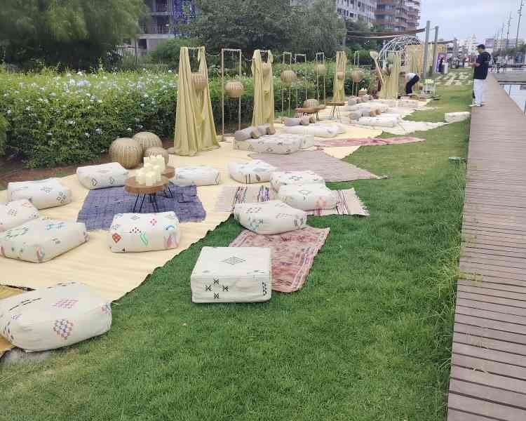 Aménagement extérieur, avec des coussins au sol, de petites tables basses et des décorations, le tout installé sur une pelouse au bord de l'eau, avec des bâtiments en arrière-plan, photo gratuite