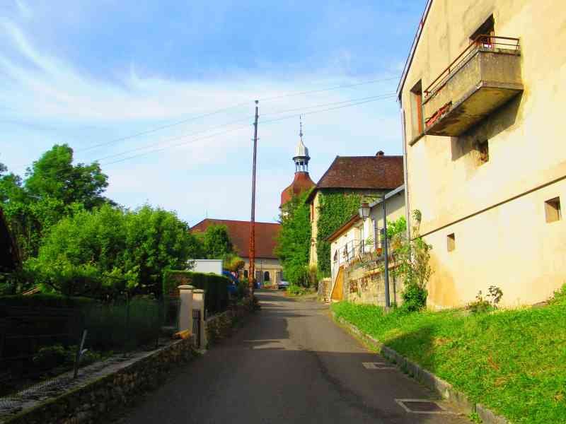Une rue de village avec des maisons traditionnelles, de la végétation et un clocher d'église visible au loin, photo gratuite
