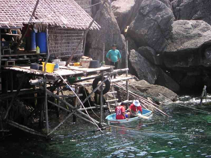 A wooden structure with a thatched roof built over the water, and people, fishermen, in a small boat below, with large rock formations in the background, free photo
